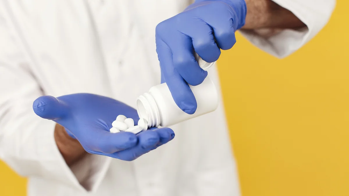 Gloved hands tipping white tablets from a medication bottle into an open palm