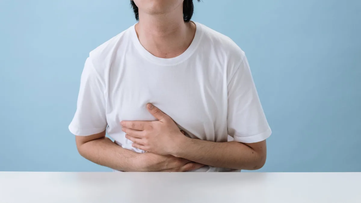Man in a white T-shirt pressing both hands against his upper abdomen at a table