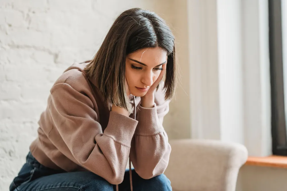 A woman sits on a sofa with her head in her hand, looking down, as in major depression